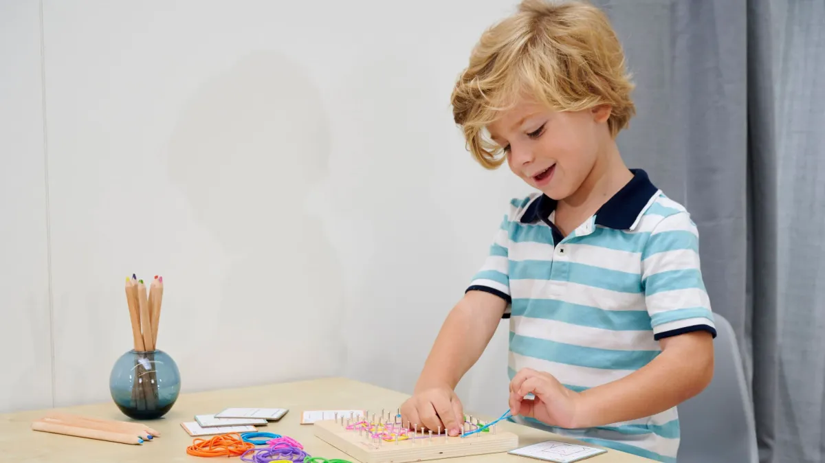 Junge spielt an einem Tisch mit einem Montessori-Geobrett und farbigen Gummibändern